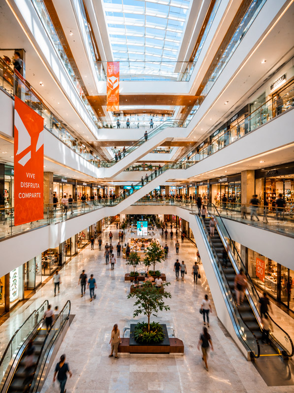IMAGEN TIPOLOGÍA CENTRO COMERCIAL: Fotografía del interior de un centro comercial moderno de varios niveles, con barandales de vidrio y acero, escaleras eléctricas, locales iluminados con vidrieras, tragaluz que aporta luz natural y acentos de color naranja en señalética. Personas comprando difuminadas. Acabados en mármol gris, blanco y madera cálida. Estilo: fotografía arquitectónica comercial vertical, realista, 4K, colores vivos y luminosos.